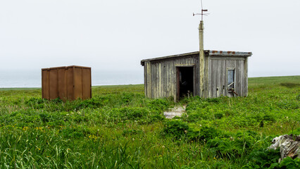 Weathered wooden cabin stands alone in a lush green field, accompanied by a rusty storage shed,...