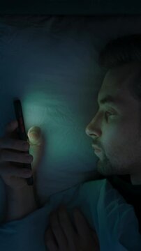 A tired man in bed scrolls endlessly on his mobile phone, struggling with insomnia, anxiety and overthinking as he watches social media late into the night. Smartphone dependence. vertical video