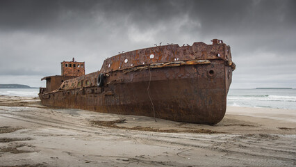Rusty shipwreck stranded on sandy beach, surrounded by turbulent waves and dark clouds, showcasing...