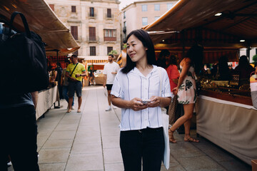 Midlife Asian woman pauses at lively street market with smartphone in hand, enjoying vibrant...