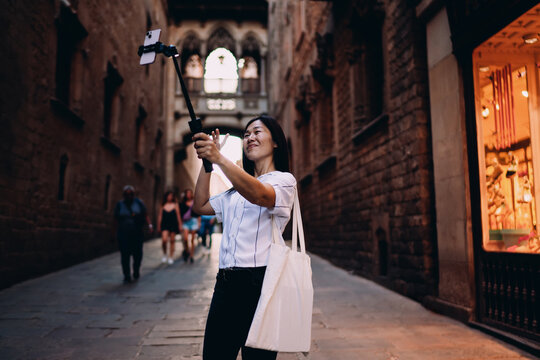 Asian woman takes selfie with stabilizer in historic street, merging travel, tech and self-representation in real-time content creation moment. - Powered by Adobe