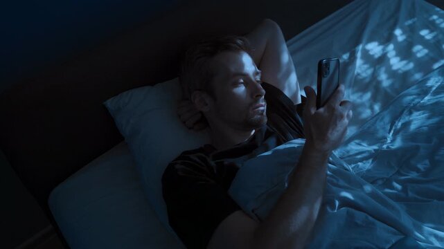 An attractive young man uses his phone late at night in bed, scrolling through his social media feed. Space for text