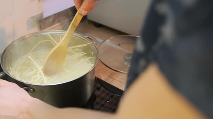 Close-up of a pot with spaghetti cooking in boiling water on a stove. A hand stirs the pasta with a wooden spoon. Home kitchen scene focused on cooking process and steam rising from the pot - Powered by Adobe