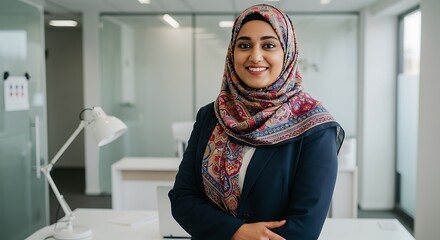 Portrait of a smiling woman in a hijab and blazer in a modern office setting looking at the camera