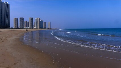 Lone coastal observer walking on urban beach with modern skyscrapers and ocean waves creating...
