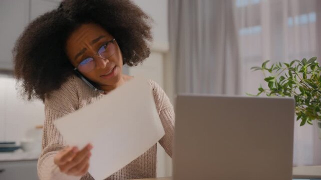 African American woman concentrated paying online with laptop and papers bills multitasking girl female business lady working laptop remote from home talking mobile phone check data in documents stats