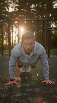 Vertical shot of young man in sport clothing doing push-ups in outdoor training area in forest at sunset