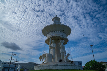 Obraz premium The sculpture at the Karon Beach roundabout in Phuket stands beneath a sky filled with small, fluffy clouds, gently diffusing the sunlight and casting a soft light over the scene. Fluffy cloud