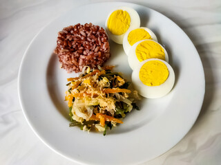 Red rice with vegetable salad or gudangan and a boiled egg on a white plate. A healthy meal concept.