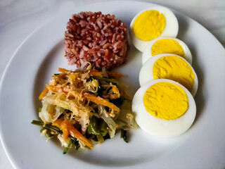 Red rice with vegetable salad or gudangan and a boiled egg on a white plate. A healthy meal concept.