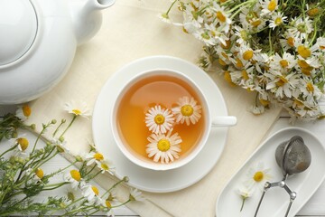 Tasty chamomile tea, infuser and flowers on white wooden table, flat lay
