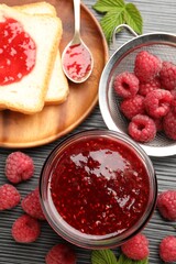Sweet raspberry jam in glass jar, berries and bread slices on dark gray wooden table, flat lay