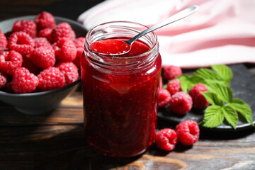 Delicious raspberry jam in glass jar, spoon, fresh berries and mint on wooden table, closeup