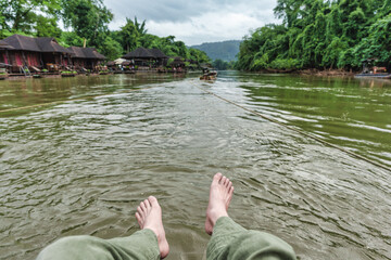 Person with feet relaxing during rafting with boat along River Kwai, lush green forest and wooden...