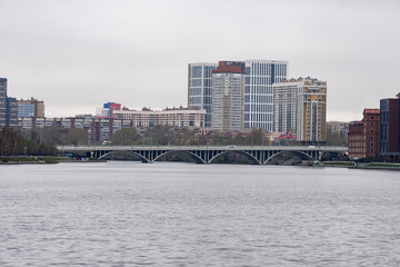 Obraz premium Panoramic view of the river in central Yekaterinburg with Bazhovsky Bridge in the distance. Cloudy sky and autumn or spring season create a moody atmosphere.
