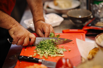 Close-up of chef’s hands chopping fresh green onions and parsley on a red cutting board with a sharp knife. Cooking preparation in a kitchen.
