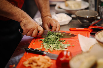 Chef chopping fresh parsley and green onions on a red cutting board with salmon and vegetables prepared on the side. Cooking process in a kitchen.