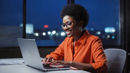 Smiling woman using laptop in office at night, with cityscape view. Her warm smile radiates joy and focus as she works