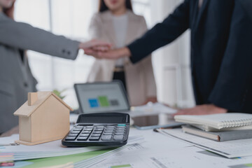 Real estate agents joining hands in celebration after closing a deal, surrounded by a house model,...