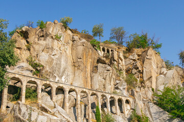 Historic stone aqueduct arches built into steep rocky cliff with trees and vegetation under clear blue summer sky