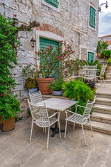 Charming outdoor cafe in a picturesque stone alley of an old Mediterranean town.Cozy restaurant terrace with white furniture and blooming flowers against a historic stone wall