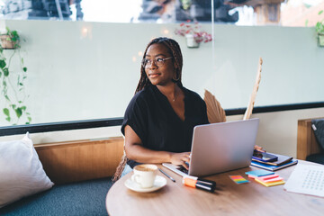 Pensive Black woman working on laptop, briefly pausing and reflecting, symbolizing thoughtful planning, digital creativity, and independent business mindset in tech-powered workspace.