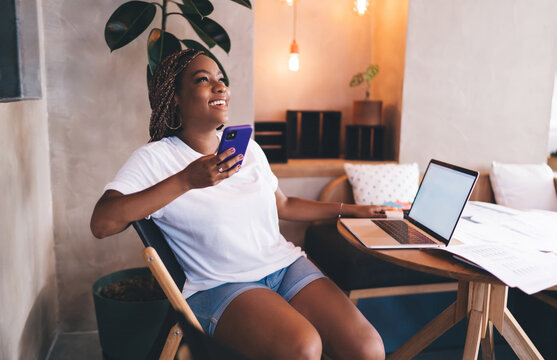 Laughing Black woman multitasks between smartphone and laptop, enjoying tech freedom and flexibility in digital work while thriving in casual, stress-free remote workflow.