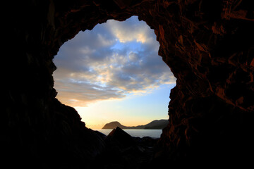 View of a cloudy sky and the ocean through a dark cave opening during a golden hour sunset time