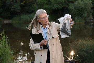 Forensic expertise. Detective with clipboard and evidence at crime scene near lake outdoors