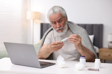 Senior man with thermometer having online consultation with doctor via laptop at table indoors