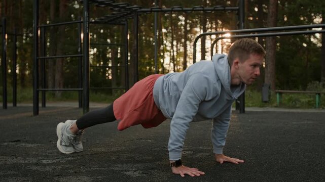Young man in sportswear doing regular push-ups as part of his calisthenics training in outdoor gym in forest at sunset