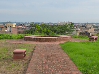 View of a scenic overlook in an urban area with a circular brick structure and pathway. Flanking the pathway are two pink stone benches on a grassy landscape.