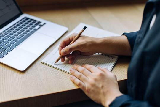 Person writing in a notebook with a laptop on a wooden desk