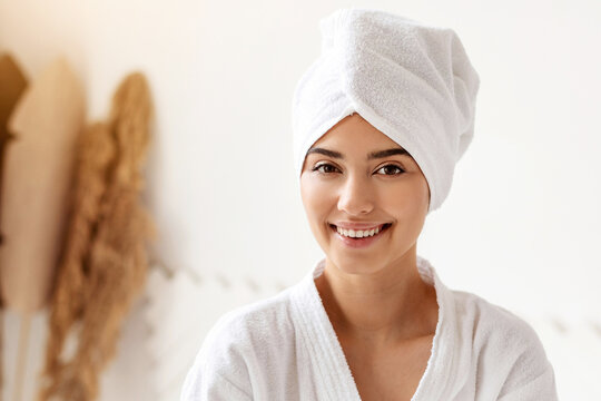 Cheerful Female Wearing White Bathrobe Posing With Towel On Head After Bathing Routine Standing In Modern Bathroom Indoor. Woman Enjoying Day At Spa Concept. Body Care And Pampering
