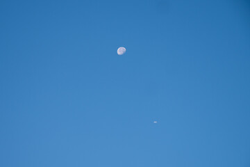 Airplane flying in clear blue sky with visible moon during daytime, minimalistic scene of aviation and astronomy.
