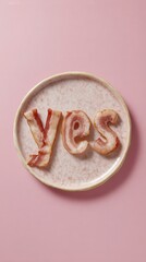 Bacon strips are creatively arranged to form the word yes on a pastel-colored plate