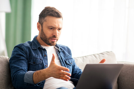 Angry middle-aged bearded man having video call with lover, having fight, using laptop, looking at computer screen, yelling and gesturing, home interior, copy space, side view