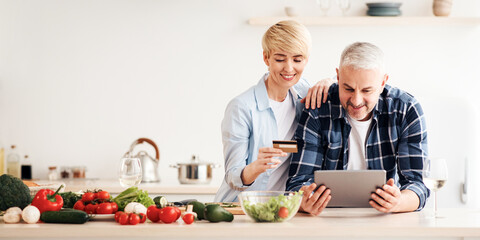 Positive emotions, shopping together, order online at home during covid-19 self-isolation. Cheerful mature couple with credit card pay money through banking on laptop at kitchen interior, empty space