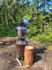 A metal kettle and samovar placed on a wooden stump in a lush green forest. Sunlight filters...