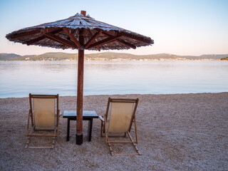 Wooden parasol with two deck chairs on the pebble beach in Biograd na Moru, Croatia, facing the calm Adriatic Sea and distant coastal town.