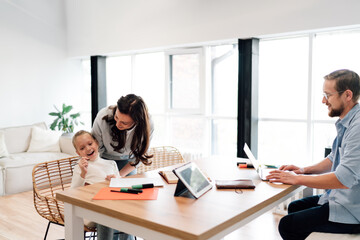 Mother assists daughter with drawing while father works on laptop, demonstrating digital harmony, multitasking lifestyle and seamless blend of work and education in tech-savvy family.