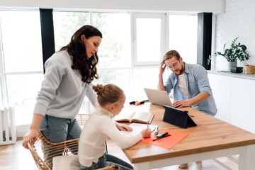 Father stressed while working on laptop, daughter focused on schoolwork, mother stands by observing, depicting pressure of multitasking and emotional intensity in tech-connected households.