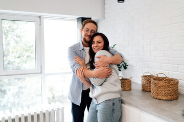 Man tenderly holds partner from behind amid kitchen’s digital hints. A relational...