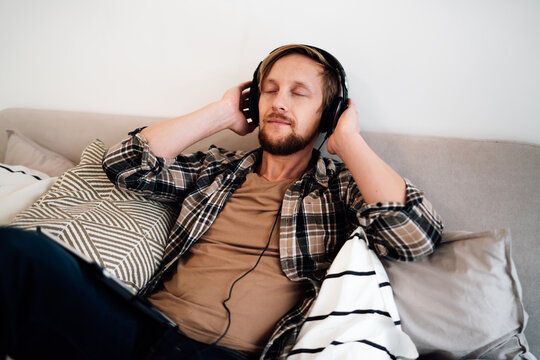 Man enjoying immersive digital moment, eyes closed with headphones on, resting on bed with tablet by side, symbolizing audio therapy, creative inspiration, and tech balance.