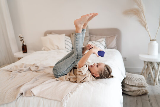 Energetic Caucasian girl lying on bed holding smartphone above, laughing while interacting with device, representing digital joy and spontaneous childhood play.