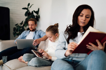 Family sits together, each absorbed in different activity—reading, using tablet, working on laptop—capturing modern multitasking routine and balanced coexistence.