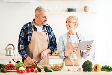 Health care in middle age, new recipe and cook together. Smiling senior male cuts vegetables, wife looks in tablet in kitchen interior with bright vegetables on table and glasses of wine in daylight