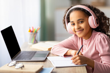 Online education concept. Happy teen girl in wireless headset using laptop and taking notes in her exercise book, smiling to camera. Computer with blank screen, mockup for ad