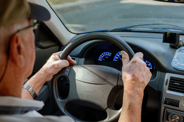 An elderly man is driving a car, holding the steering wheel with both hands, and has vitiligo on...