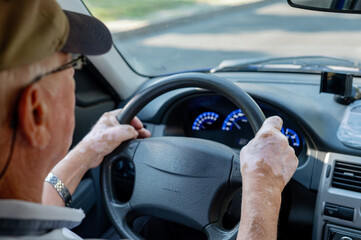 An elderly man is driving a car, holding the steering wheel with both hands, and has vitiligo on...